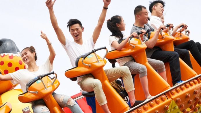 six people sitting on a orange ride and holding onto black handles, one man is cheering with his arms in the air and another woman has her thumbs up