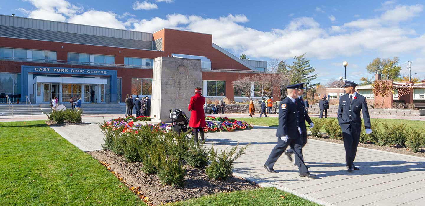 three men dressed in suits walking by person in red coat looking at statue in front of the East york civic centre