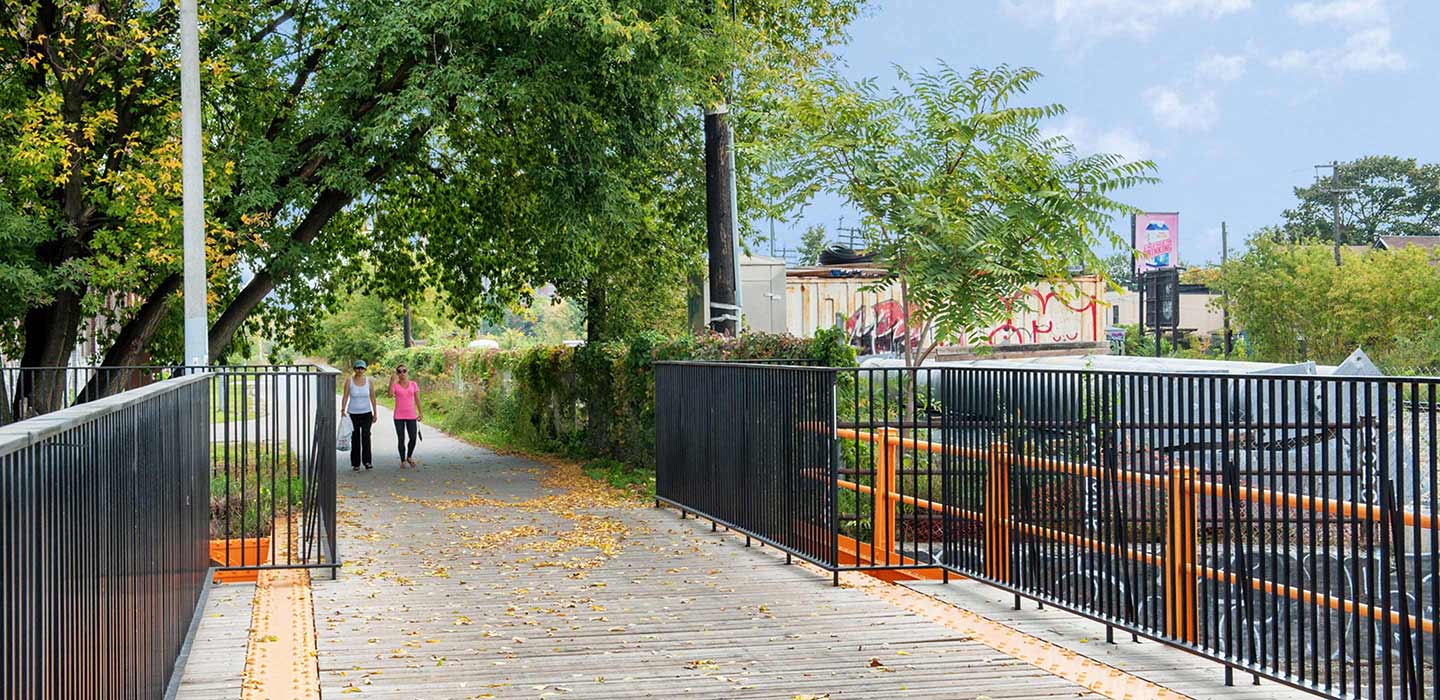 two women walking on boardwalk lined with black fences and greenery at West Toronto Railpath