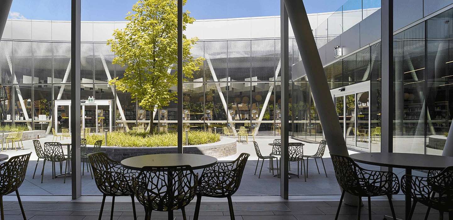 Looking out large windows of the Vaughn Civic Centre to centre outdoor courtyard with tables and chairs and a large green tree in the centre